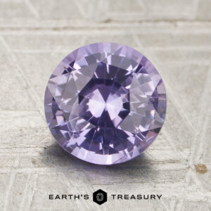 A 2.02-carat, round-cut, faceted lavender sapphire (heated) is displayed on a textured surface with the text “Earth’s Treasury” below.