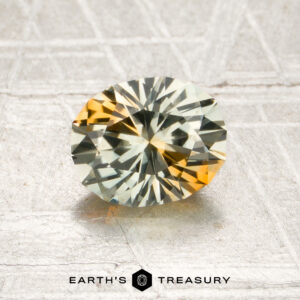 A 1.33-carat near colorless-orange particolored Montana sapphire, cut in a round faceted shape, displays pale yellow and orange hues as it rests on a textured silver surface. The “Earth’s Treasury” logo appears at the bottom of the image.