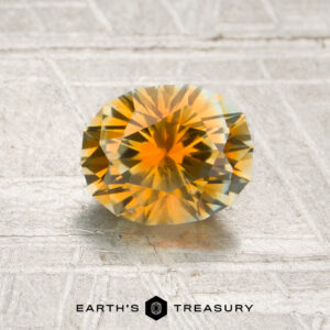 A 1.29-carat orange Montana sapphire (heated), faceted in a round shape, is displayed on a textured, light-colored surface with the “Earth’s Treasury” logo below.