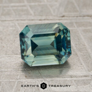 A 2.05-carat teal green Madagascar sapphire (heated) in a rectangular emerald cut is displayed on a textured gray surface, with the Earth’s Treasury logo at the bottom.
