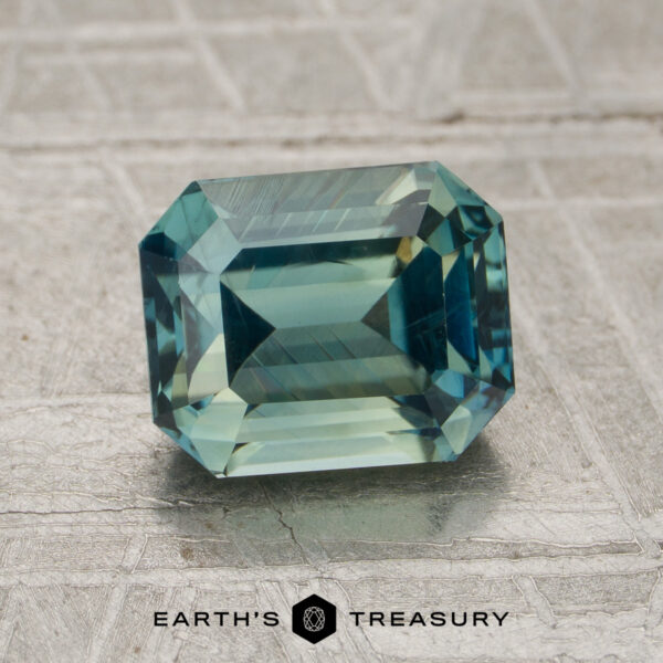 2.05-Carat Teal Green Madagascar Sapphire (Heated) A 2.05-carat teal green Madagascar sapphire (heated) in a rectangular emerald cut is displayed on a textured gray surface, with the Earth’s Treasury logo at the bottom.
