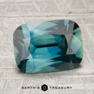 A rectangular, faceted 2.56-carat teal Madagascar sapphire (heated) rests on a textured gray surface with the words “EARTH’S TREASURY” below.
