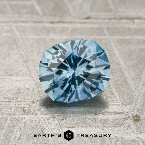 1.18-Carat Aqua Blue Montana Sapphire (Heated) A 1.18-carat heated Montana sapphire with a faceted blue appearance rests on a textured surface, with the words “EARTH’S TREASURY” and a hexagon logo displayed below.
