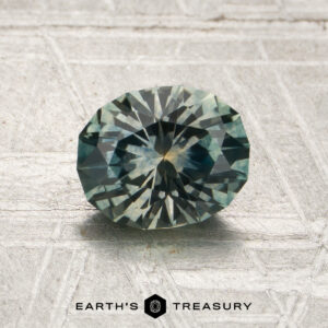 A 1.25-carat gray-green Montana sapphire (heated) in an oval faceted cut rests on a patterned surface, with the Earth’s Treasury logo displayed below.