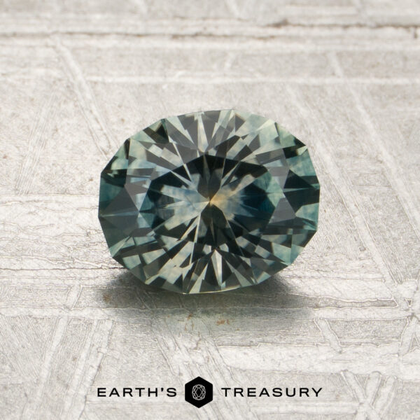 1.25-Carat Gray-Green Montana Sapphire (Heated) A 1.25-carat gray-green Montana sapphire (heated) in an oval faceted cut rests on a patterned surface, with the Earth’s Treasury logo displayed below.