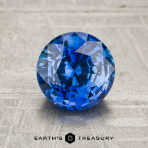 A 1.91-carat rich blue Ceylon sapphire (heated) with intricate facets is displayed on a textured light surface, with the text “EARTH’S TREASURY” beneath the stone.