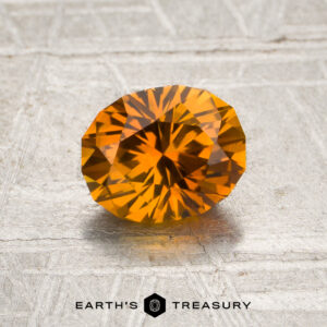 A 1.25-carat rich orange Montana sapphire (heated) is displayed on a textured light surface, with the text “EARTH’S TREASURY” and a hexagon logo below it.