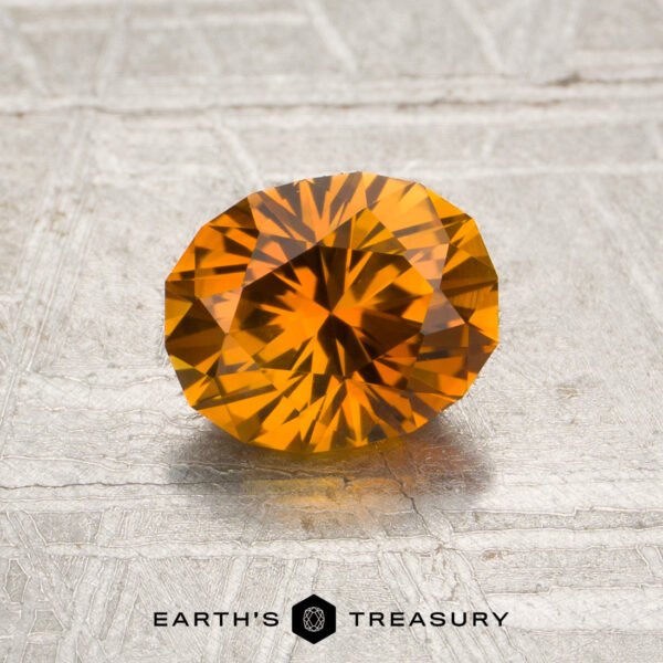 1.25-Carat Rich Orange Montana Sapphire (Heated) A 1.25-carat rich orange Montana sapphire (heated) is displayed on a textured light surface, with the text “EARTH’S TREASURY” and a hexagon logo below it.