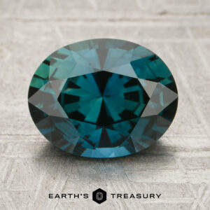 A 2.69-carat deep teal Madagascar sapphire (heated), expertly faceted in an oval shape, rests on a textured gray surface with the “EARTH’S TREASURY” logo and text below.