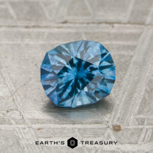 A 1.13-carat heated Montana sapphire with a vivid blue faceted cut rests on a textured metallic surface, displaying the Earth’s Treasury logo and text at the bottom.