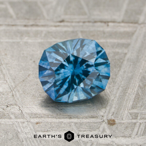 1.13-Carat Teal Blue Montana Sapphire (Heated) A 1.13-carat heated Montana sapphire with a vivid blue faceted cut rests on a textured metallic surface, displaying the Earth’s Treasury logo and text at the bottom.