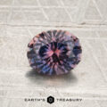 A 1.19-carat Violet-Lavender-Orange particolored Montana sapphire, round and faceted, rests on a textured gray surface. The Earth’s Treasury logo is visible at the bottom of the image.