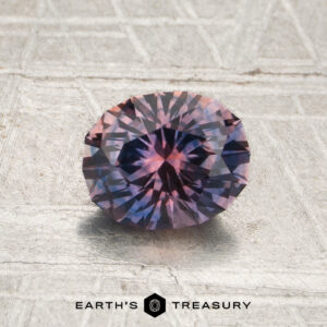A 1.19-carat Violet-Lavender-Orange particolored Montana sapphire, round and faceted, rests on a textured gray surface. The Earth’s Treasury logo is visible at the bottom of the image.