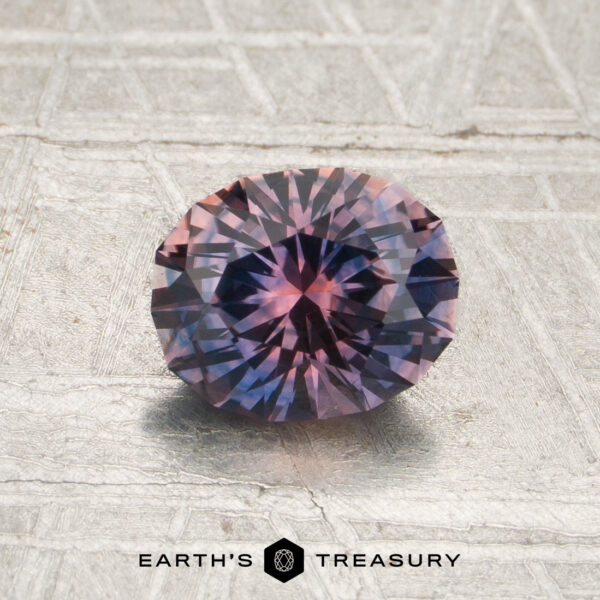 1.19-Carat Violet-Pink-Orange Particolored Montana Sapphire (Heated) A 1.19-carat Violet-Lavender-Orange particolored Montana sapphire, round and faceted, rests on a textured gray surface. The Earth’s Treasury logo is visible at the bottom of the image.