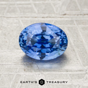 A 1.67-carat unheated oval-cut blue sapphire is displayed on a textured metallic surface, with the Earth’s Treasury logo at the bottom.