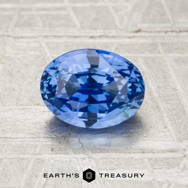 1.67-Carat Cornflower Blue Ceylon Sapphire (Unheated) A 1.67-carat unheated oval-cut blue sapphire is displayed on a textured metallic surface, with the Earth’s Treasury logo at the bottom.
