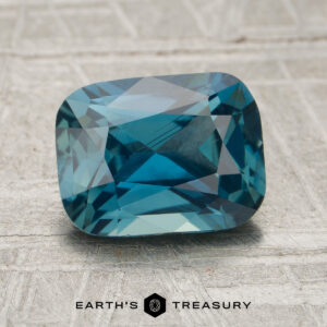 A 2.26-carat teal Madagascar sapphire, cut in a rectangular shape, is displayed on a textured surface with the “Earth’s Treasury” logo beneath it.