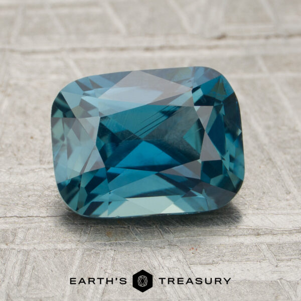 2.26-Carat Teal Madagascar Sapphire (Unheated) A 2.26-carat teal Madagascar sapphire, cut in a rectangular shape, is displayed on a textured surface with the “Earth’s Treasury” logo beneath it.