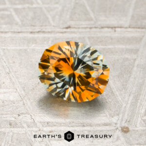 1.14-carat orange-near colorless particolored Montana sapphire with an oval cut, displaying vibrant orange and green hues on a textured surface, accompanied by the Earth’s Treasury logo and text below.