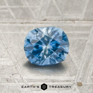 A 1.12-carat heated Montana sapphire is displayed on a textured gray surface with the words “EARTH’S TREASURY” below it.