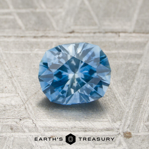 A 1.12-carat heated Montana sapphire is displayed on a textured gray surface with the words “EARTH’S TREASURY” below it.