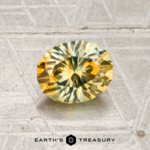 A 1.13-carat mint green-orange particolored Montana sapphire (heated) with an oval cut, displayed on a textured metallic surface and the words “EARTH’S TREASURY” at the bottom.