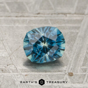 A 1.08-carat heated Montana sapphire is displayed on a textured, metallic surface with the text “EARTH’S TREASURY” and a hexagonal logo beneath it.
