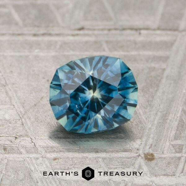 1.08-Carat Teal Montana Sapphire (Heated) A 1.08-carat heated Montana sapphire is displayed on a textured, metallic surface with the text “EARTH’S TREASURY” and a hexagonal logo beneath it.