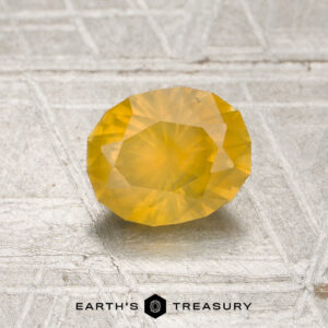 A 1.10-carat opalescent golden yellow Montana sapphire (heated) rests on a textured surface, with the words “EARTH’S TREASURY” and a logo displayed below.