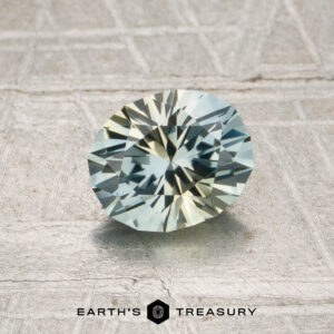A 1.07-carat pale blue-yellow particolored Montana sapphire (heated), cut in a round brilliant style, rests on a textured surface; the “Earth’s Treasury” logo appears at the bottom.