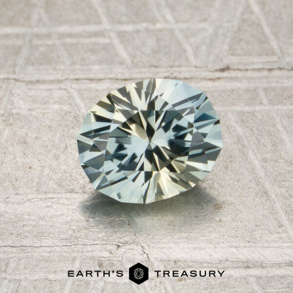 1.07-Carat Pale Blue-Yellow Particolored Montana Sapphire (Heated) A 1.07-carat pale blue-yellow particolored Montana sapphire (heated), cut in a round brilliant style, rests on a textured surface; the “Earth’s Treasury” logo appears at the bottom.