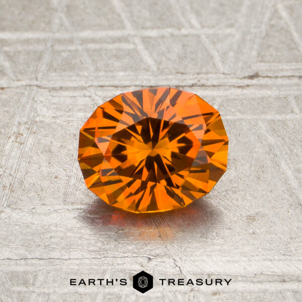 1.03-Carat Intense Orange Montana Sapphire (Heated) A 1.03-carat intense orange Montana sapphire (heated), expertly oval-cut, is showcased on a textured silver surface, accompanied by the text “EARTH’S TREASURY” and a hexagonal logo beneath.