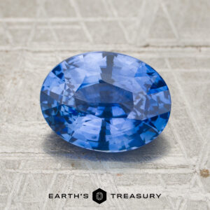 A 3.55-carat cornflower blue to violet color-change Ceylon sapphire, oval-shaped, displayed on a textured gray surface with the “EARTH’S TREASURY” logo and text beneath it.