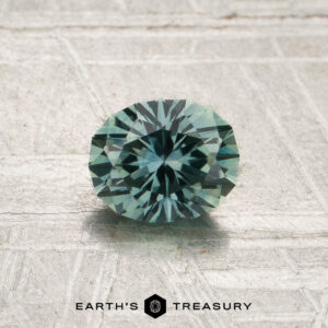 A 0.90-carat spruce green Montana sapphire (heated), expertly cut in a round brilliant shape, is displayed on a textured light surface. The logo and text “Earth’s Treasury” appear at the bottom of the image.