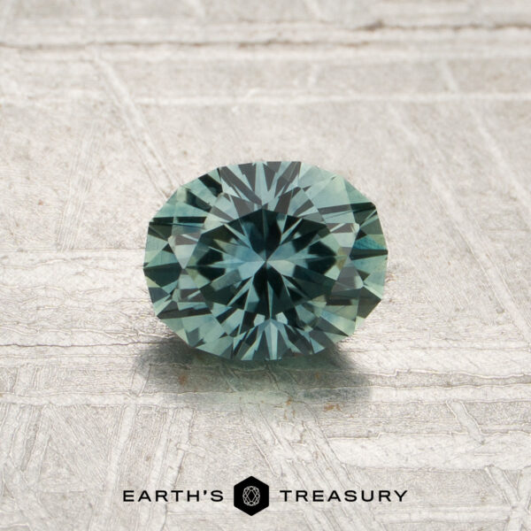 0.90-Carat Spruce Green Montana Sapphire (Heated) A 0.90-carat spruce green Montana sapphire (heated), expertly cut in a round brilliant shape, is displayed on a textured light surface. The logo and text “Earth’s Treasury” appear at the bottom of the image.
