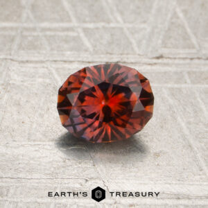 A faceted oval 0.88-carat orange-red Montana sapphire (heated) rests on a textured gray surface, with the text “EARTH’S TREASURY” and a hexagon logo at the bottom.