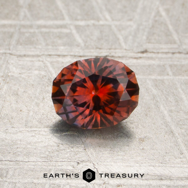 0.88-Carat Orange-Red Montana Sapphire (Heated) A faceted oval 0.88-carat orange-red Montana sapphire (heated) rests on a textured gray surface, with the text “EARTH’S TREASURY” and a hexagon logo at the bottom.