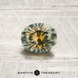 A 0.85-carat mint green-orange particolored Montana sapphire (heated) is displayed on a textured surface with the text “EARTH’S TREASURY” at the bottom.