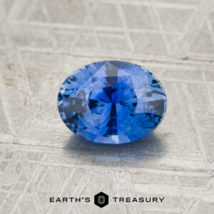 A 2.30-carat cornflower blue Ceylon sapphire gemstone rests on a textured silver surface, featuring the “Earth’s Treasury” text and logo at the bottom of the image.