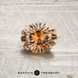A 0.85-carat peach-orange particolored Montana sapphire (heated), cut in a round brilliant style, is displayed on a textured surface with the “Earth’s Treasury” logo and text visible at the bottom of the image.