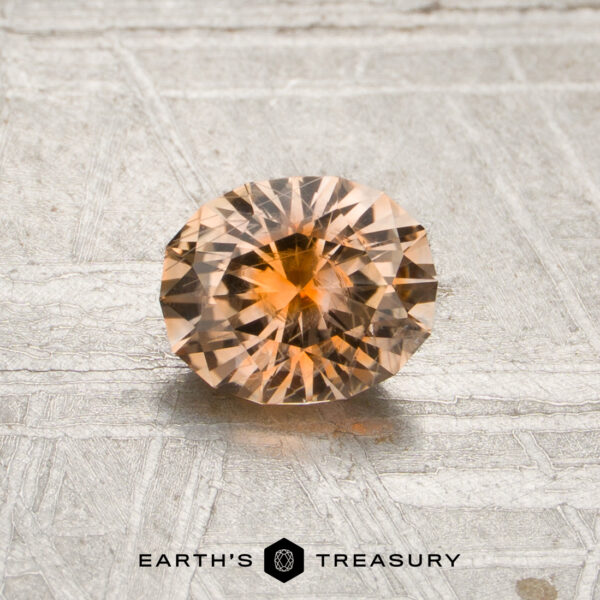 0.85-Carat Peach-Orange Particolored Montana Sapphire (Heated) A 0.85-carat peach-orange particolored Montana sapphire (heated), cut in a round brilliant style, is displayed on a textured surface with the “Earth’s Treasury” logo and text visible at the bottom of the image.