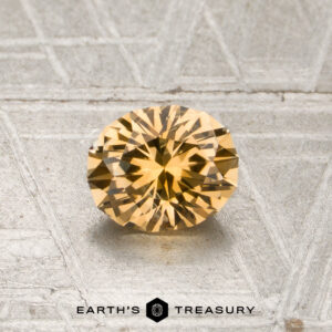 A round, faceted 0.85-carat light orange Montana sapphire (heated) displayed on a textured, light gray surface with the “Earth’s Treasury” logo and text at the bottom.
