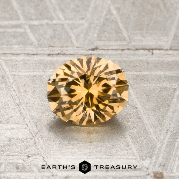 0.85-Carat Light Orange Montana Sapphire (Heated) A round, faceted 0.85-carat light orange Montana sapphire (heated) displayed on a textured, light gray surface with the “Earth’s Treasury” logo and text at the bottom.