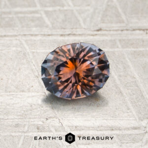 A 0.77-carat orange-purple particolored Montana sapphire (heated), cut in a faceted oval shape, is displayed on a textured light surface with the text “Earth’s Treasury” at the bottom.