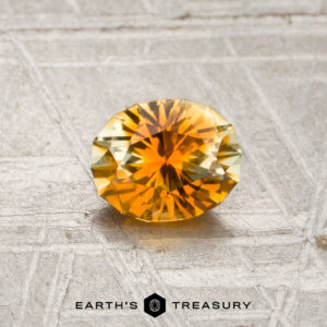 A 0.76-carat rich orange-colorless particolored Montana sapphire is displayed on a textured gray surface above the text “EARTH’S TREASURY.”.