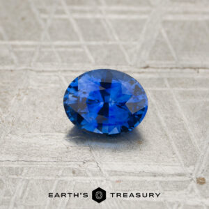A 1.19-carat rich blue to violet color-change Ceylon sapphire is displayed on a textured, metallic surface, with “EARTH’S TREASURY” appearing at the bottom.
