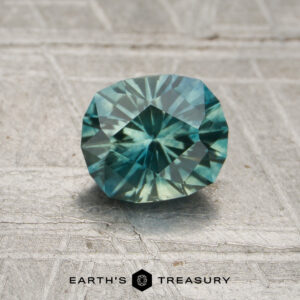 A 0.96-carat Montana sapphire (heated), showcasing vibrant blue-green facets, rests on a textured gray surface above the “EARTH’S TREASURY” text and a hexagonal logo.