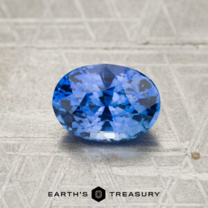 A 2.23-carat Cornflower Blue to Violet color-change Ceylon sapphire, expertly cut in an oval shape, rests on a textured gray surface with the Earth’s Treasury logo at the bottom.