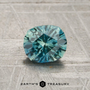 A 0.91-carat Montana Sapphire (heated), featuring a blue-green faceted cut, is showcased on a textured gray surface with the Earth’s Treasury logo and name displayed at the bottom.