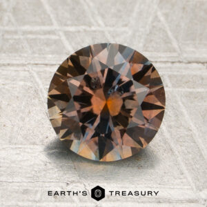 A 1.94-carat orange-pale blue particolored Montana sapphire (heated), cut in a round shape, rests on a textured surface with the text “EARTH’S TREASURY” displayed below the stone.